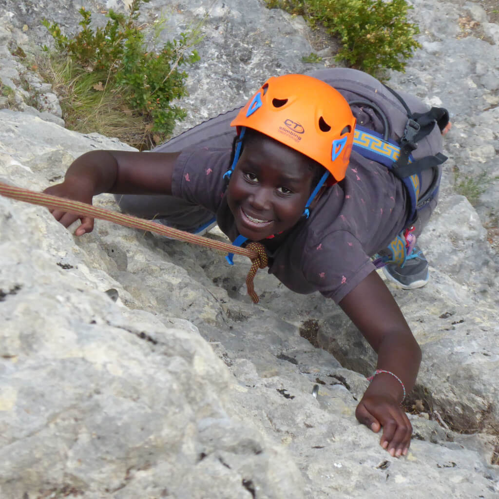 Une jeune personne portant un casque d'escalade et un harnais orange vif escalade une falaise rocheuse dans la région de la Drôme, en France. Elle s'agrippe à la roche avec ses mains et ses pieds, utilisant le matériel d'escalade pour faciliter son ascension.