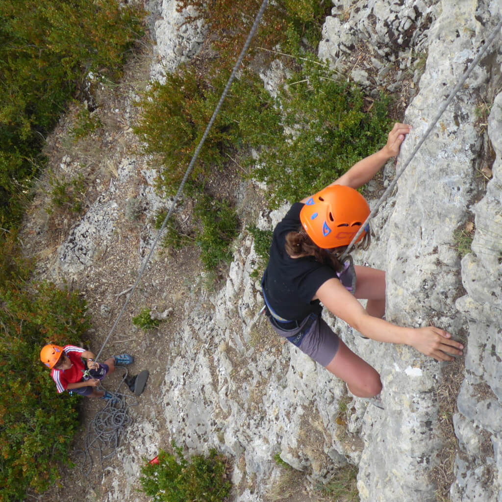 Une jeune fille escalade une falaise rocheuse escarpée dans la Drôme, utilisant ses mains et ses pieds pour agripper la surface inégale. Elle porte un casque orange vif et de l'équipement d'escalade pour l'aider dans son ascension.